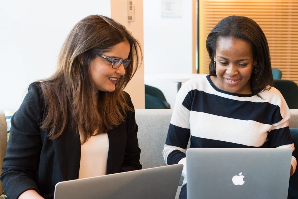 Two women dressed in business casual attire sit together in front of two open laptops.