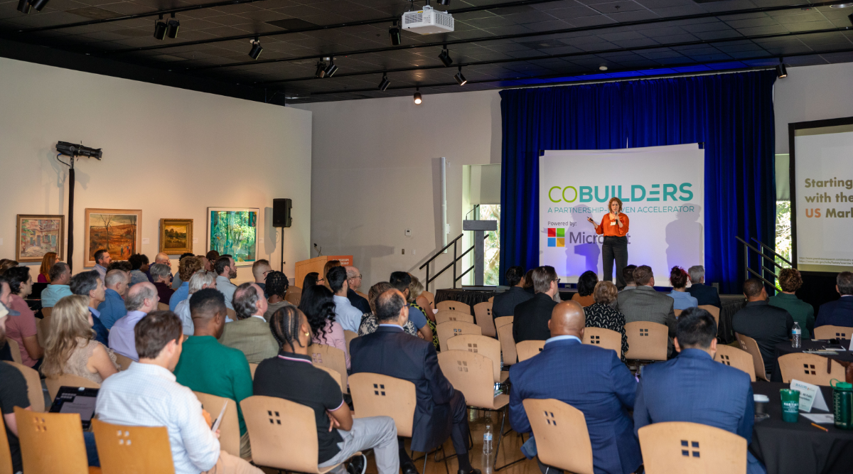 Several people seated in rows watching a woman present on a stage at the front of the room with a CoBuilders backdrop